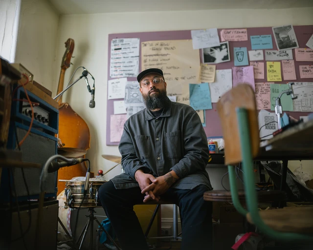 A man with a beard wearing glasses, a hat, and a blue shirt sits in a room surrounded by instruments and poster art.