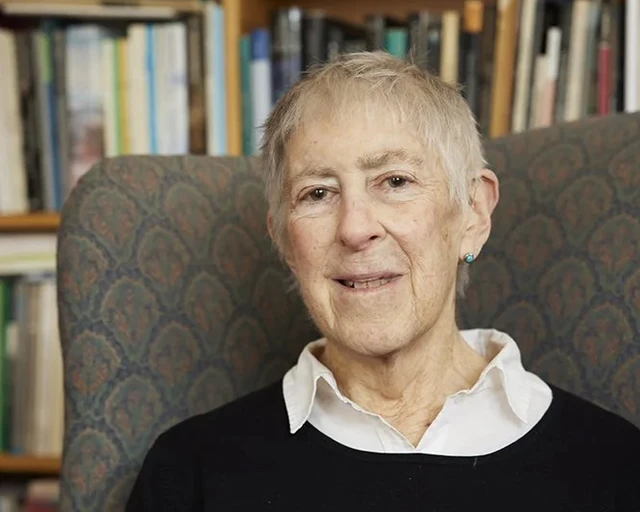 A white woman with short grey hair wearing a black sweater over a white collared shirt sits in a grey chair in front of a bookshelf.