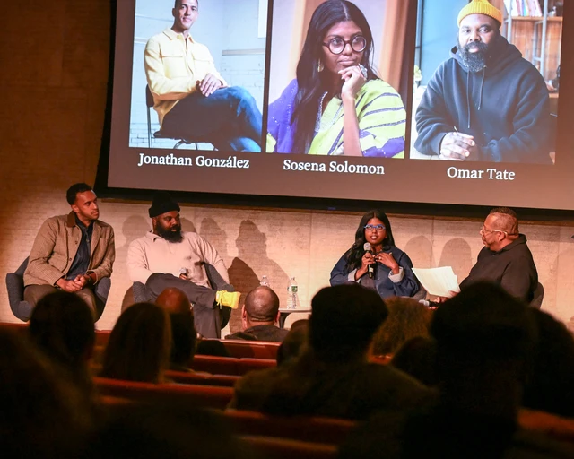 The image shows a panel discussion taking place in a dimly lit event space. Four individuals are seated on stage, engaged in conversation. Behind them is a large screen displaying photos and names of Jonathan González, Sosena Solomon, and Omar Tate. An audience is visible in the foreground.