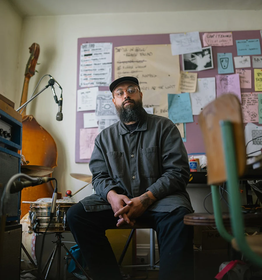 A man with a beard wearing glasses, a hat, and a blue shirt sits in a room surrounded by instruments and poster art.